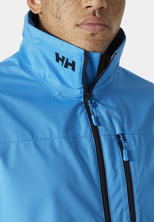 Chaqueta azul clara con un alto cuello, cremallera negra y un sutil detalle del logo. Textura suave y bolsillos prácticos para almacenamiento.