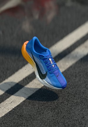 Blå og gule Nike løbesko i luften over en asfaltvej med hvide linjer, hvor de puffer små partikler op.