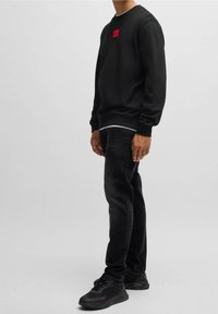 Sudadera negra con escote redondo y un pequeño parche de logo rojo en el pecho, combinada con vaqueros oscuros y zapatillas negras.