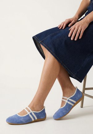 Kvinde sidder på en stol iført blå ruskinds Mary Jane-flats med hvide remme og brune såler, kombineret med en mørk denimnederdel.