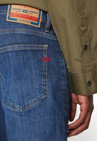 Denim jeans with a dark blue wash, featuring a red embroidered logo on the pocket and a tan leather label. Straight leg design.
