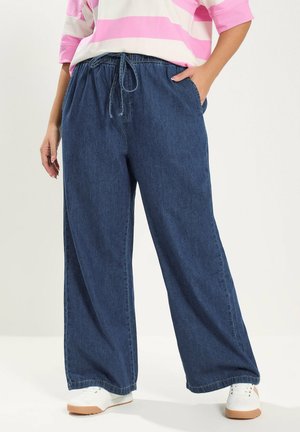 Person trägt blaue weite Jeanshosen mit Kordelzugbund, kombiniert mit weißen Sneakers und einem rosa-weiß gestreiften Oberteil.