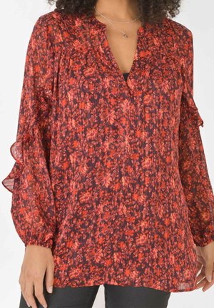 Kobieta nosząca czerwoną i pomarańczową kwiecistą bluzkę z długim rękawem, lekkimi falbankami na rękawach i dekoltem w serek, zestawioną z czarnymi spodniami.