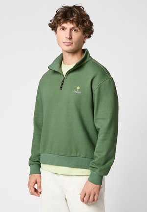 Jeune homme portant un sweat-shirt vert à fermeture éclair quart, avec le logo FAGUO, une chemise vert clair en dessous, et un pantalon blanc sur un fond clair uni.