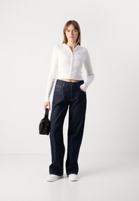Top bianco a coste con zip abbinato a jeans a gamba larga blu scuro. L'outfit è completato da una piccola borsa nera e scarpe bianche.