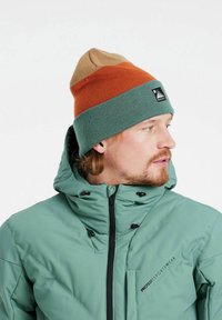 Gorra de punto con un diseño de tres colores: beige, naranja y azul verdoso. Se lleva con una chaqueta acolchada azul verdoso con cremallera frontal y capucha.