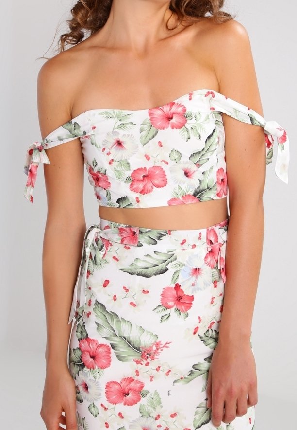 Femme portant une tenue deux pièces blanche à fleurs avec un haut court épaules dénudées et une jupe taille haute assortie aux motifs floraux roses et verts.