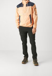 Chaqueta con capucha en color durazno y azul marino, con un bolsillo frontal y detalles con cremallera, combinada con pantalones negros y zapatos de senderismo marrones.