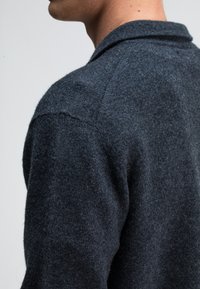 Dunkelgrauer Wollpullover mit strukturiertem Finish, ausgestattet mit einem breiten Kragen und taillierten Schultern, zur Seitenansicht vor einem hellen Hintergrund.