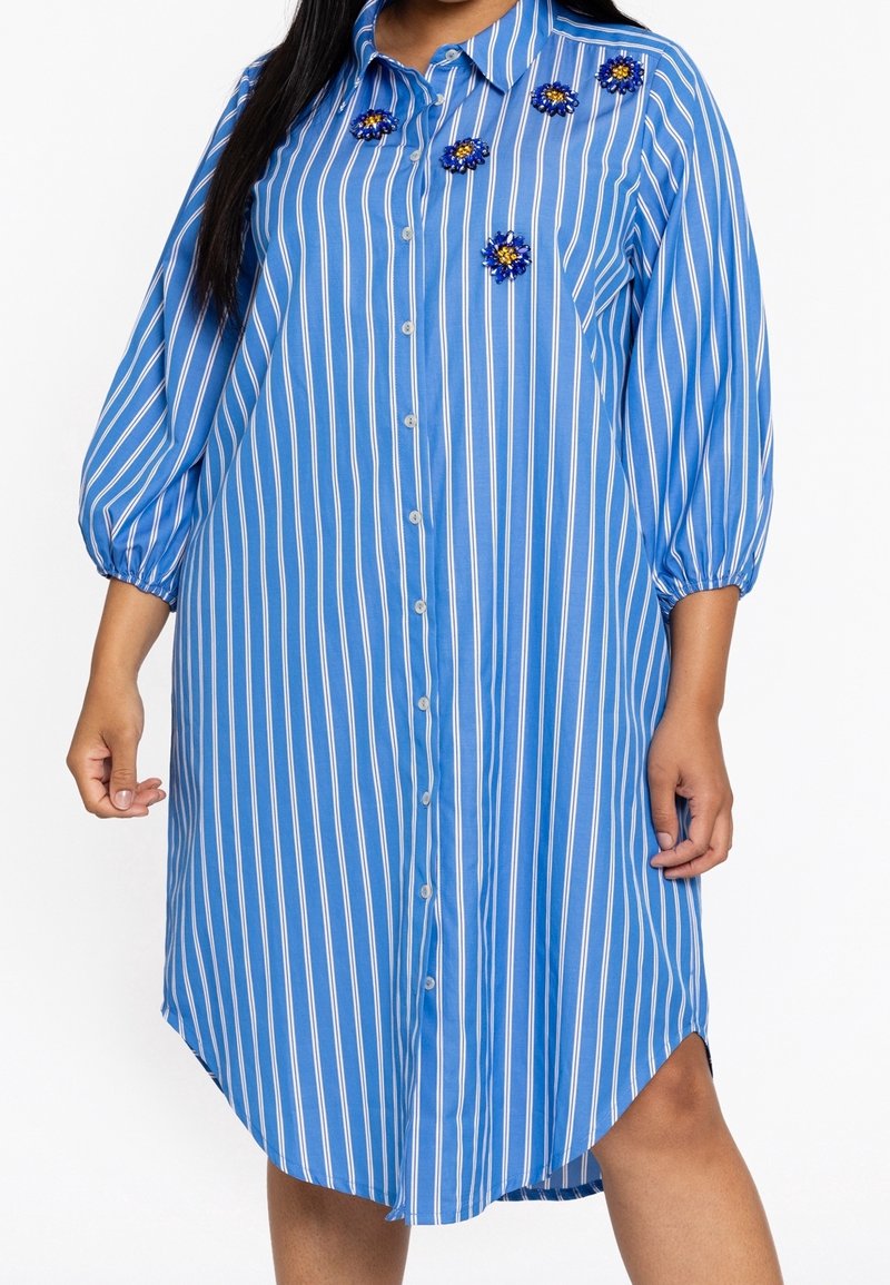 Femme portant une robe chemise rayée bleu et blanc à motifs verticaux avec des manches trois-quarts et des ornements en forme de fleurs près du col.