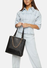 Svart läder tote-väska med texturerade accenter, guld kedjedetalj och vinklad design, hålls av en person i en denimoutfit.
