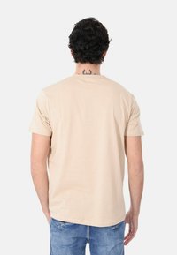 Maglietta beige a maniche corte con scollo rotondo e texture liscia, indossata con jeans azzurri chiari. La vista posteriore non mostra grafiche o loghi.