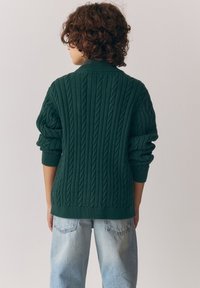 Tamsiai žalias kabelinės megztinio megztinis su aukštu apykakliu ir susuktomis rankovėmis, derinamas su šviesiai mėlynais laisvos formos džinsais, matomas iš nugaros.