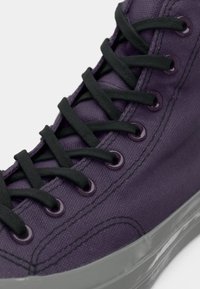 Purpursarkanva skeiters ar melnām auklām, caurumiem un šuvēm. Izteikta caurspīdīga pelēka gumijas zole. Tuva skata augšējā daļa.