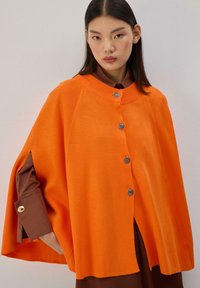 Cape orange tricotée avec un col rond, ornée de boutons argentés et de manches larges, portée par-dessus une chemise brune. Design texturé et vibrant.