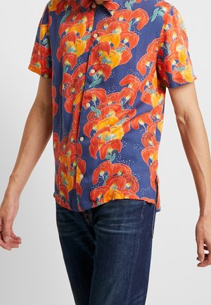 Mand iført en kortærmet blå skjorte med store røde og orange blomsterprint og mørkeblå jeans.