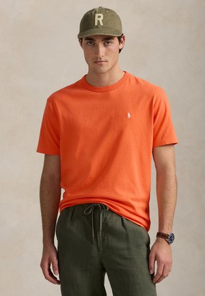 Jonge man die een oranje T-shirt met korte mouwen, olijfgroene broek, olijfgroene pet met de letter "R" en een horloge draagt, tegen een effen achtergrond.