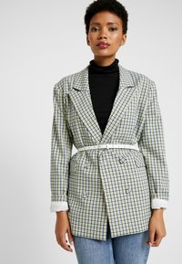 Blazer a scacchi in verde, blu e bianco. Design strutturato con rever ampi, chiusura doppio petto e una cintura bianca. Tessuto leggero.