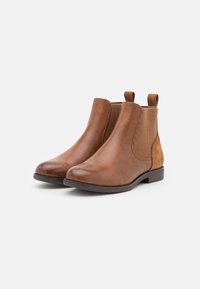 Bottines en cuir marron avec des panneaux latéraux en elastique et un accent texturé en beige. Bout arrondi et talon bas. Surface lisse avec un minimum de détails.