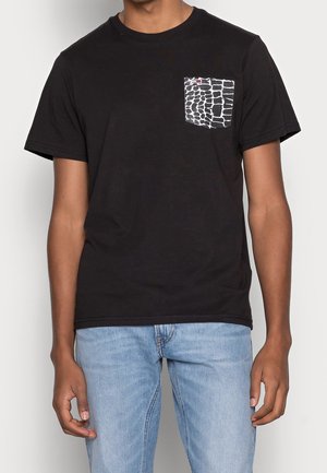Mann trägt ein schwarzes T-Shirt mit kurzen Ärmeln und einer weiß-schwarzen gemusterten Brusttasche, kombiniert mit hellblauen Jeans.