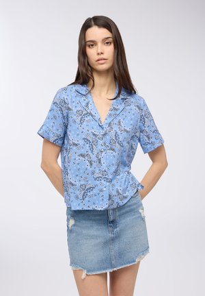 Blusa azzurro chiaro a maniche corte con colletto, caratterizzata da un motivo paisley nero. Abbinata a una mini gonna di jeans strappata.