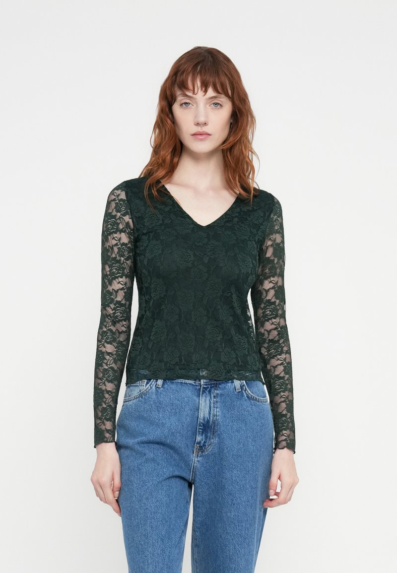 Haut vert en dentelle à manches longues avec un décolleté en V, orné de motifs floraux. Associé à un jean en denim bleu taille haute.