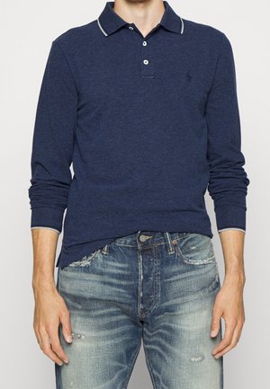 Marineblaue Langarm-Polo-Shirt mit Kragen, zwei Knöpfen und kontrastierenden Akzenten am Kragen. Getragen mit verwaschenen Jeans.