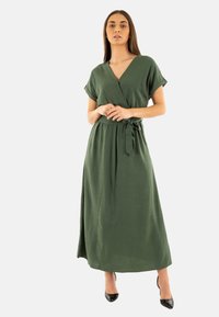 Vestido verde de largo hasta la rodilla, tipo envoltura, con mangas cortas y una cintura ajustada con cinta. Tiene un escote en forma de V y una textura de tela suave. Combinado con tacones negros.