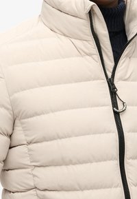 Giacca a vento beige chiaro con cuciture orizzontali, colletto alto e zip completa con un tenditore nero e accento con logo. Tessuto morbido.