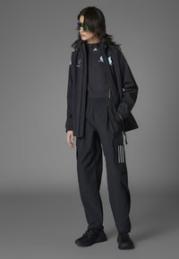 Sortt sportsoutfit bestående af en jakke med lynlås, en tætsiddende overdel og løse bukser med cargo-lommer og tre-stribede detaljer. Mørke sneakers fuldender looket.