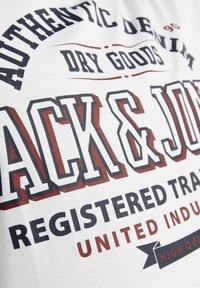 Hvit T-skjorte med stor flerfarget trykt tekst: "AUTENTISK DENIM," "TØRRE VARER," og "JACK & JONES" med dekorative detaljer.