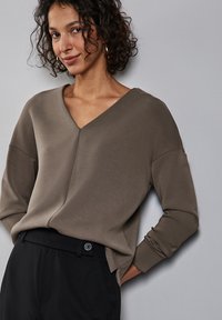 Felpa marrone a V con spalle scese e maniche lunghe, realizzata in tessuto morbido. Indossata con pantaloni neri sartoriali.