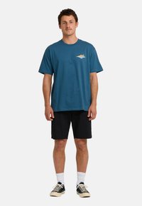 Mann steht in einem blauen Grafik-T-Shirt, schwarzen Shorts, weißen Socken und schwarzen Turnschuhen vor einem hellen einfarbigen Hintergrund.