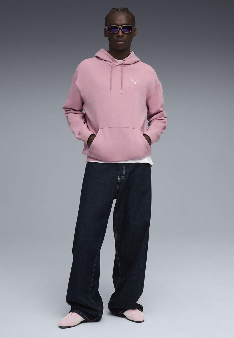 Felpa rosa con cappuccio, tasca anteriore e logo bianco, abbinata a jeans larghi blu scuro e sneakers rosa chiaro.