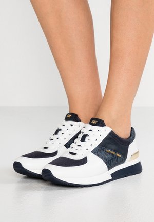 Osoba nosząca białe i granatowe sneakersy Michael Kors ze złotym logo i akcentami, stojąca na gładkiej jasnej podłodze.