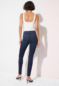 Jeans ajustados de denim oscuro con cintura alta y dos bolsillos traseros, combinados con una blusa sin mangas blanca y mules negros de tacón.