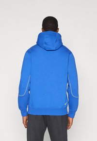 Sudadera azul con una gran capucha, costuras amarillas de acento a lo largo de los lados y puños acanalados. La tela parece suave y cómoda.