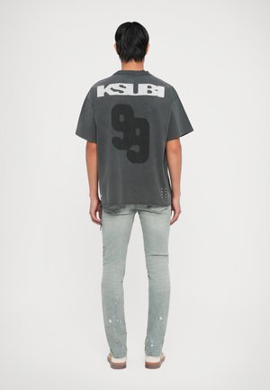 Gray oversized T-shirt with "KSUBI" text, large black graphic, short sleeves, paired with light wash jeans featuring paint splatters.