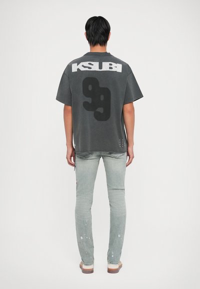 Graues, übergroßes T-Shirt mit "KSUBI"-Schriftzug, großem schwarzen Grafikdesign, kurzen Ärmeln, kombiniert mit hellblauen Jeans mit Farbspritzern.