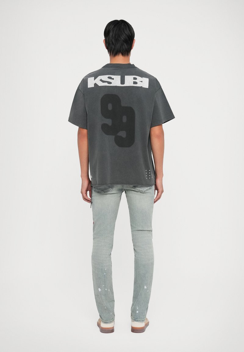 Grijze oversized T-shirt met de tekst "KSUBI", grote zwarte graphic, korte mouwen, gecombineerd met licht gewassen jeans met verfspetters.
