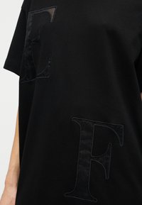 Μαύρο υπερμέγεθες μπλουζάκι από βαμβάκι, με μεγάλα υφασμάτινα γράμματα 'E' και 'F' σε ταιριαστό μαύρο ύφασμα, που δημιουργούν μια διακριτική αντίθεση σχεδίου.