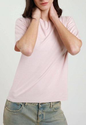 Donna con maglietta a maniche corte rosa chiaro che si tiene il collo con entrambe le mani, indossando jeans blu slavati su uno sfondo semplice.