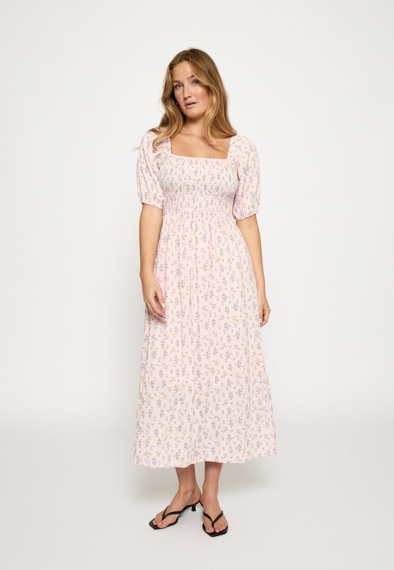 Moteris dėvi rožinės gėlėtos vidutinio ilgio suknelę su pūstomis rankovėmis ir kvadratine iškirpte, stovinti atsipalaidavusia poza priešais vienodą foną.