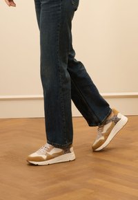 Witte en beige sneakers met leoprintaccenten, vetersluiting, dikke zool, gedragen met donkerblauwe opgerolde jeans op een houten vloer.