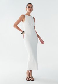 Robe maxi blanche à côtes avec une silhouette cintrée, un décolleté rond et une légère évasée à l'ourlet. Des tongs noires complètent le look.