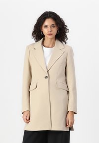 Manteau beige ajusté avec un col à revers, fermeture à un bouton, poches avant et forme structurée, fabriqué en tissu texturé.