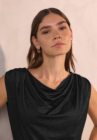 Zwarte, mouwloze top met gedrapeerde halslijn, gemaakt van een glanzende, gestructureerde stof. Model draagt gouden oorringen.