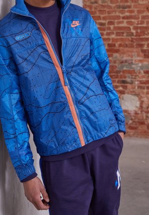 Homme portant une veste Nike Air bleue avec un design de carte, une fermeture éclair orange et un pantalon de survêtement Nike bleu foncé, debout contre un mur en briques.