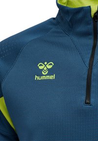 Giacca sportiva blu con colletto a mezzo zip, tessuto strutturato e dettagli verdi. Presenta il logo "hummel" in verde brillante sul petto.