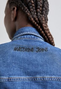 Traper jakna s ovratnikom, tamnim šavovima i crnim "Moschino Jeans" logotipom na leđima. Tekstura se čini glatkom i istrošenom.